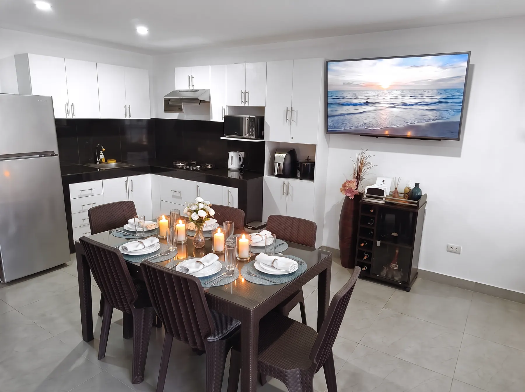 Comedor para seis servido con vajilla blanca y velas, junto a la cocina y la TV de 55 pulgadas