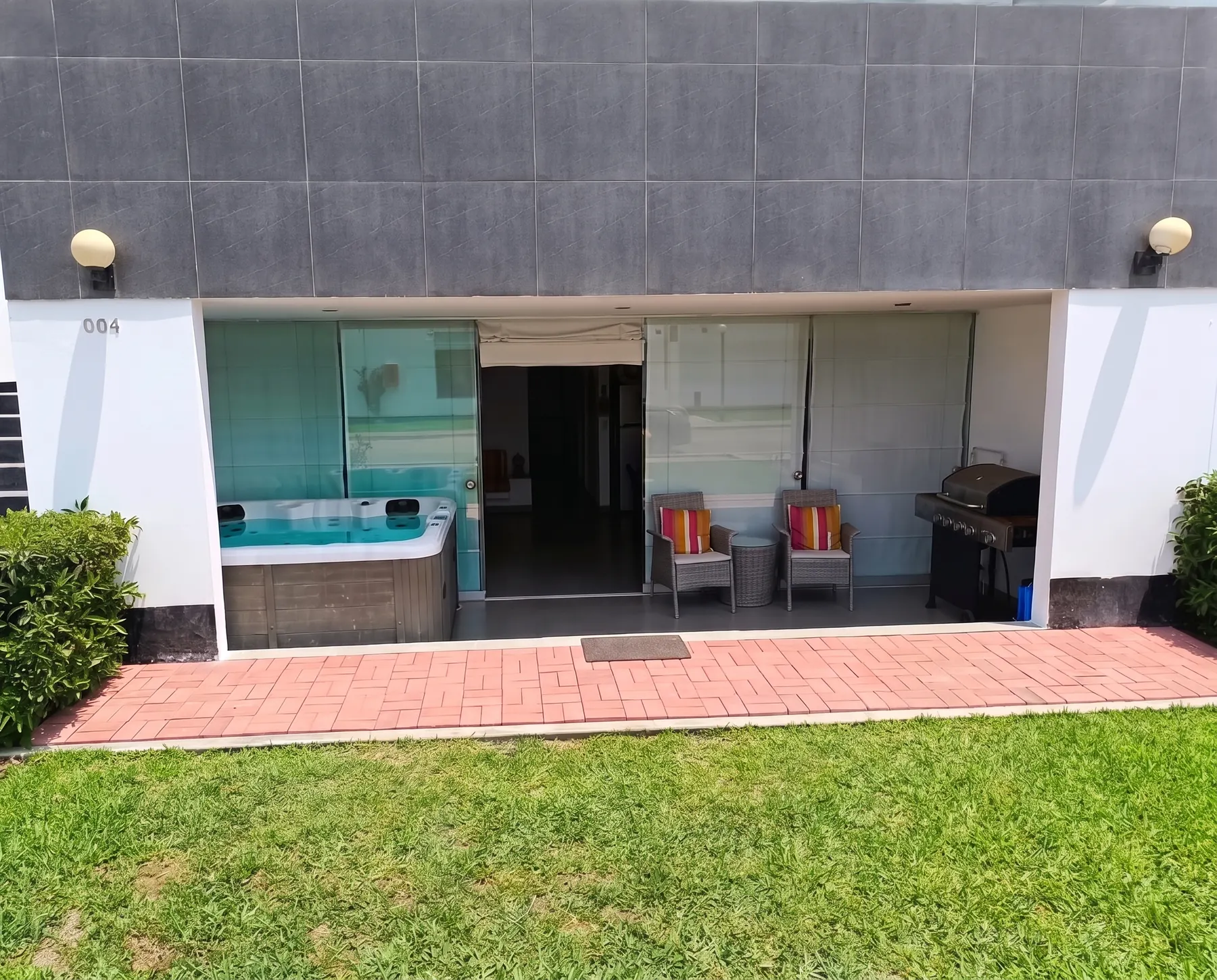 Fachada del depa de día: terraza techada con jacuzzi, sillones de mimbre y parrilla frente al jardín
